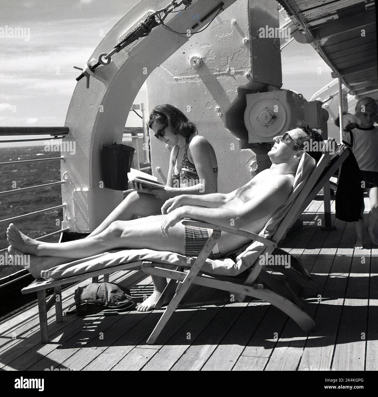 Late 1960s, historical, happy cruising, an adult couple in their bathing suits relaxing in deckchairs on the deck of the Norweigian-America cruise ship, MS Sagafjord. The male passenger is lying getting some sun, while the lady is reading a book. Stock Photo