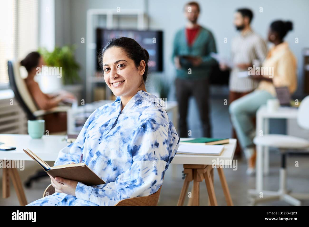 Happy young businesswoman with open book sitting by workplace against