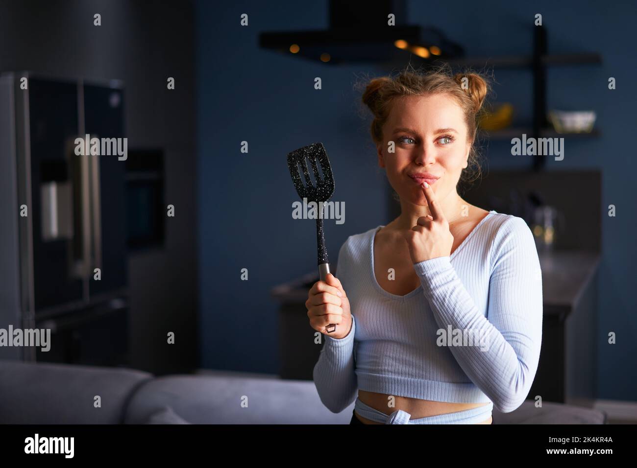 Young thoughtful caucasian blonde woman holding black spatula smiling cunningly standing at home on a dark modern stylish kitchen background. Young Stock Photo