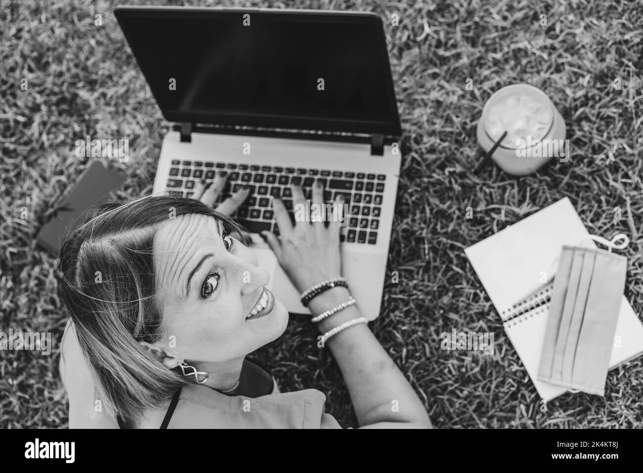 Woman 40 years old working outdoors with laptop while making healthy breakfast - Smiling female entrepreneur or influencer lying down on lawn typing o Stock Photo