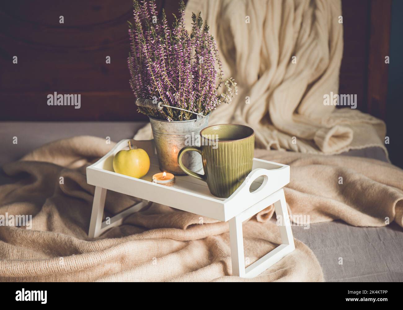 Cute white wooden breakfast tray with legs on home bed with cup of hot tea, tea light candle and heather flower. Autumn arrangement set with scarf. Stock Photo