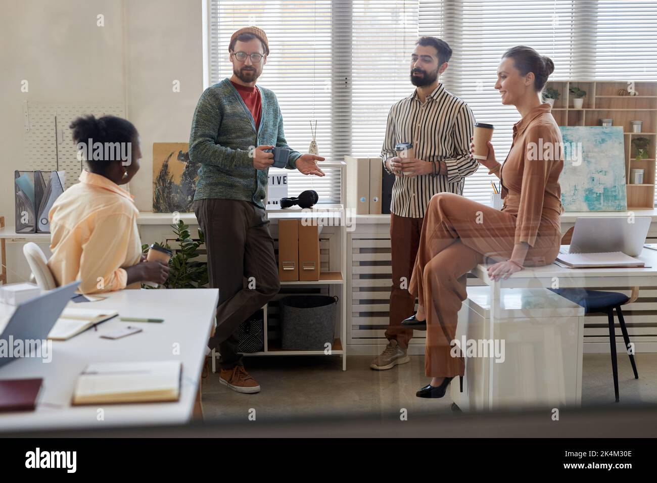 Four young multicultural colleagues with cups of coffee discussing news while businessmen standing in front of two female colleagues Stock Photo