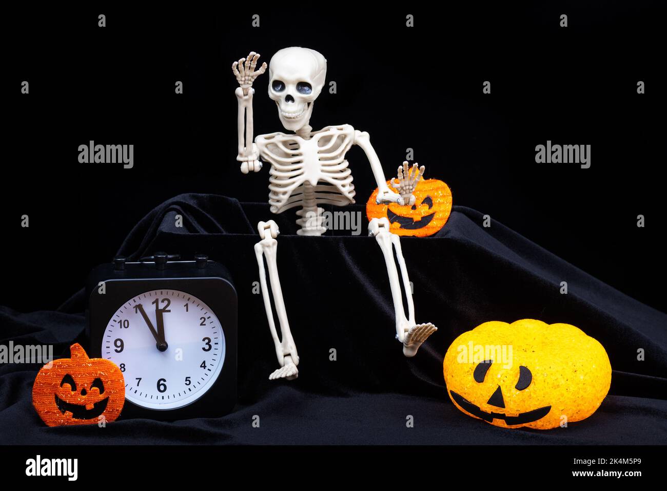 Halloween time. A skeleton crawling out of the grave and waving his hand, greeting, pumpkins with grimaces, a clock on a black background. The eve of Stock Photo