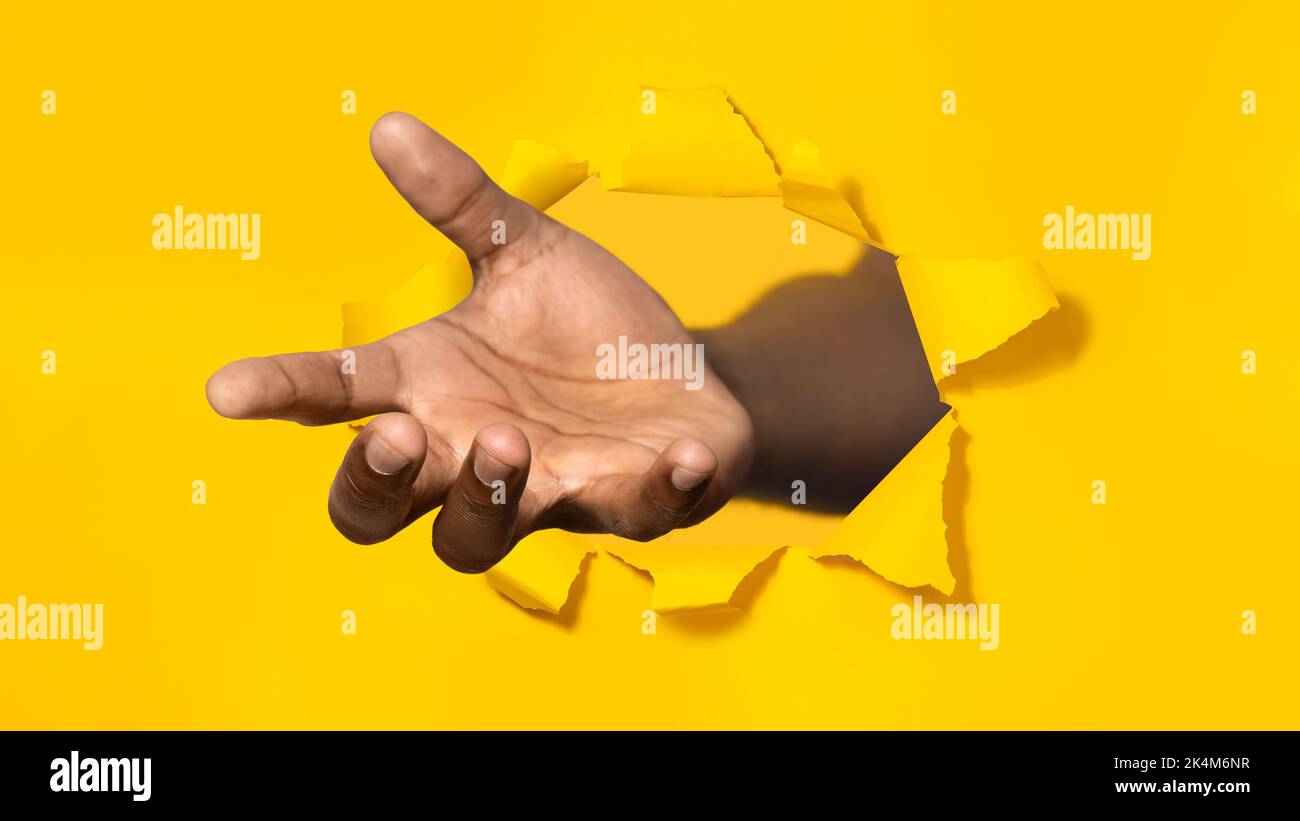 African american man stretching open palm through hole in torn yellow paper, offering helping hand, asking for charity Stock Photo