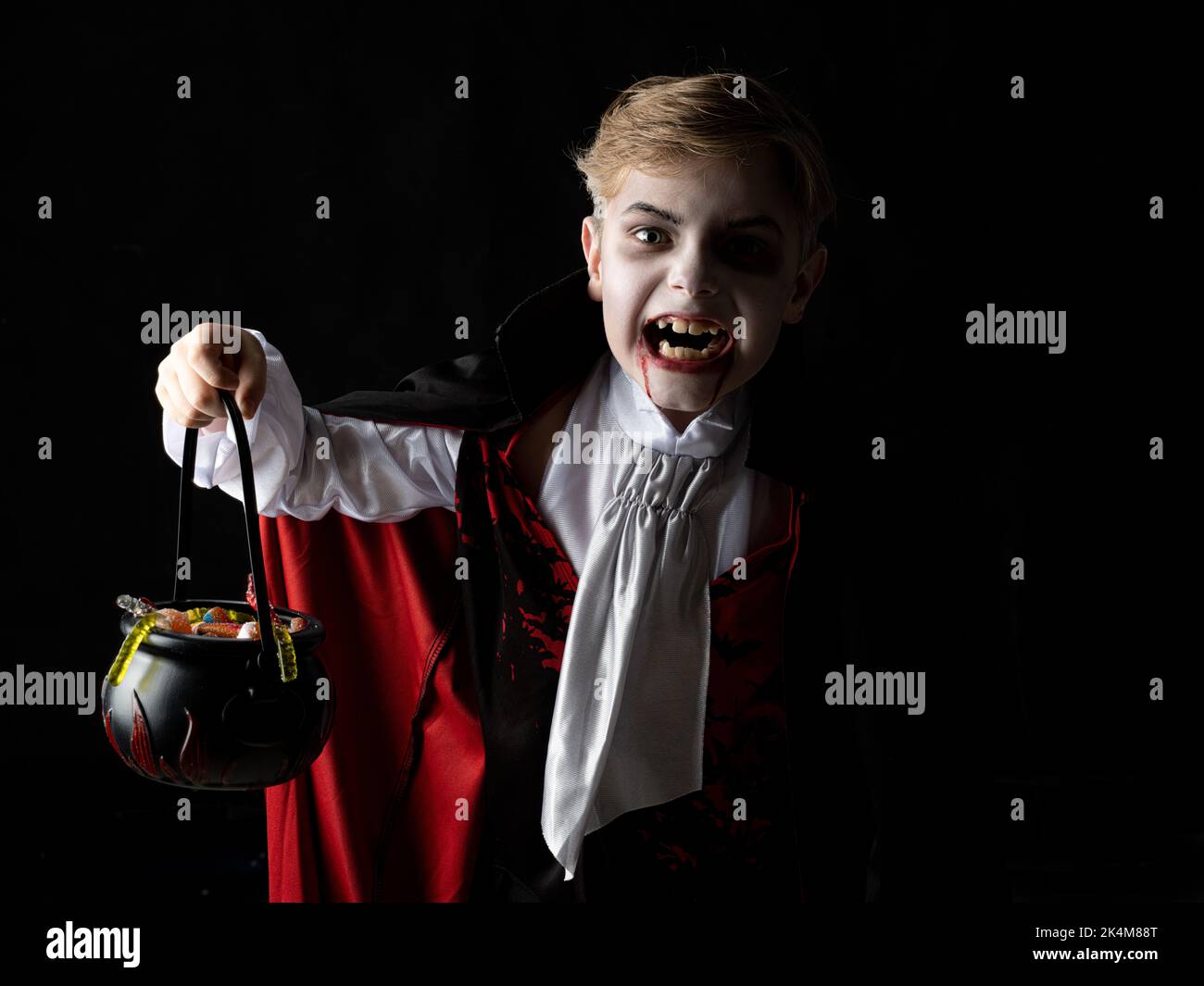 Happy child boy in costume of vampire holding cauldron od candies on black background in holiday Halloween Stock Photo