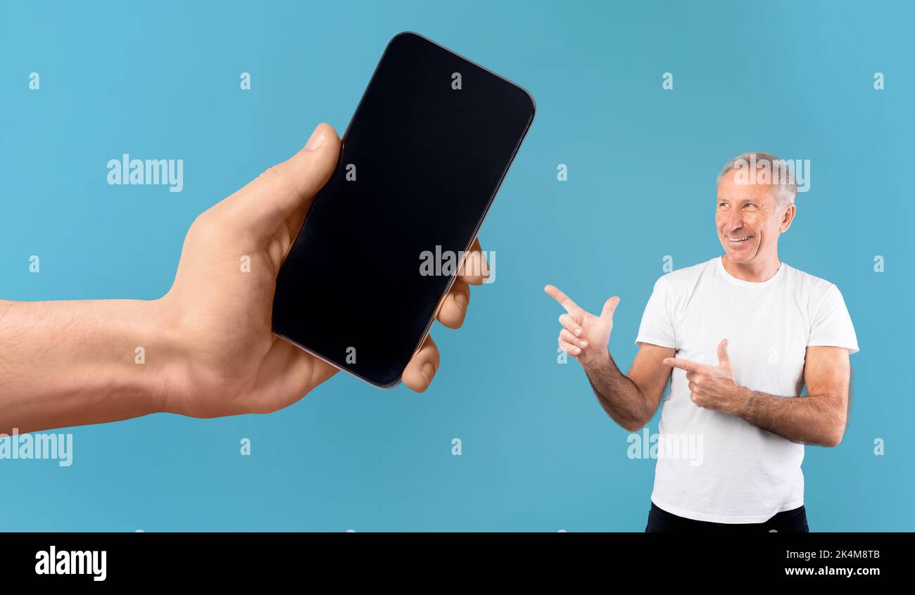 Cheerful elderly european male pointing fingers at huge smartphone with blank screen in hand Stock Photo