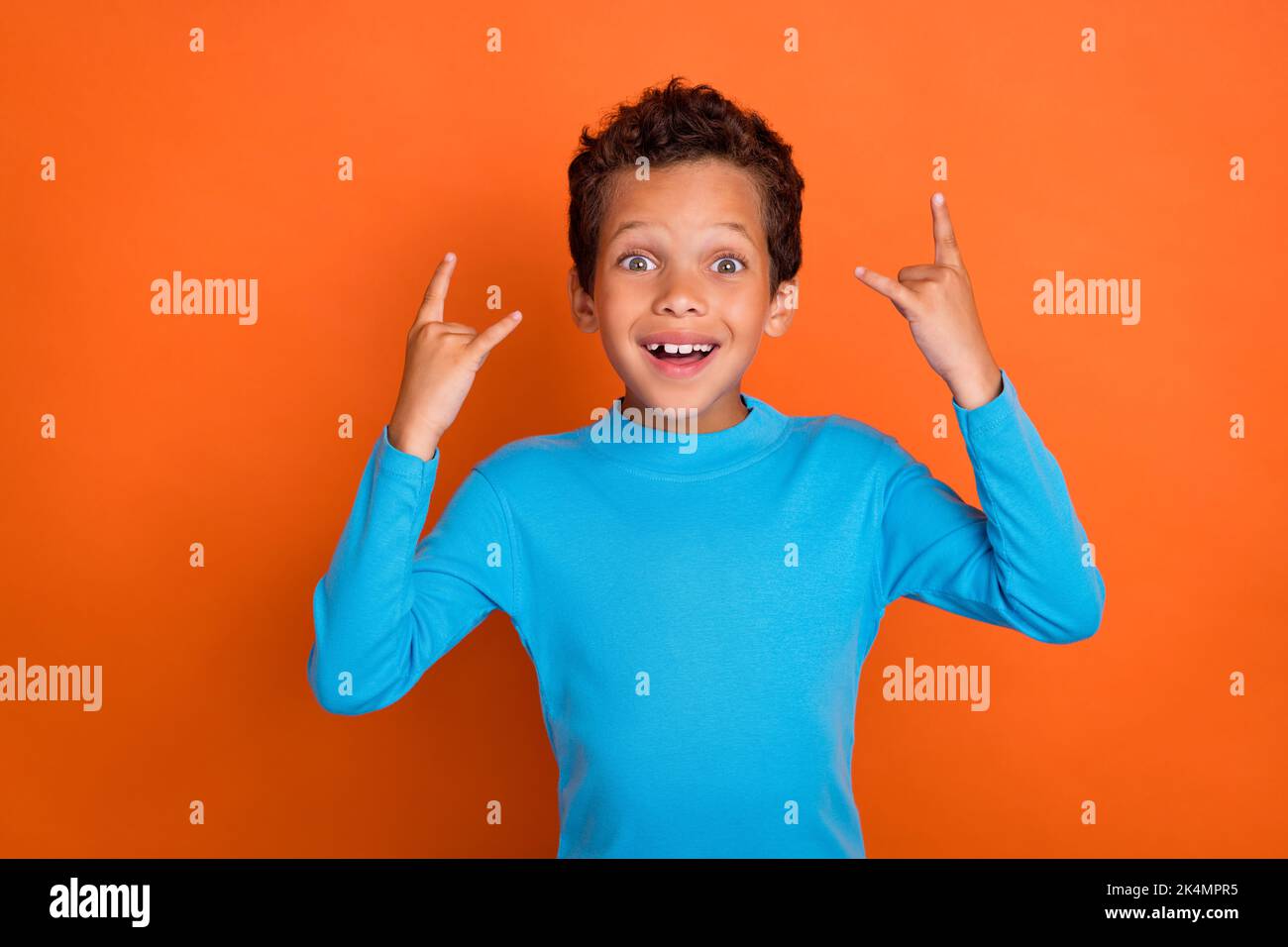 Photo of cheerful boy fan rock music arm demonstrates symbol toothy smile dressed blue trendy sweater isolated on orange color background Stock Photo