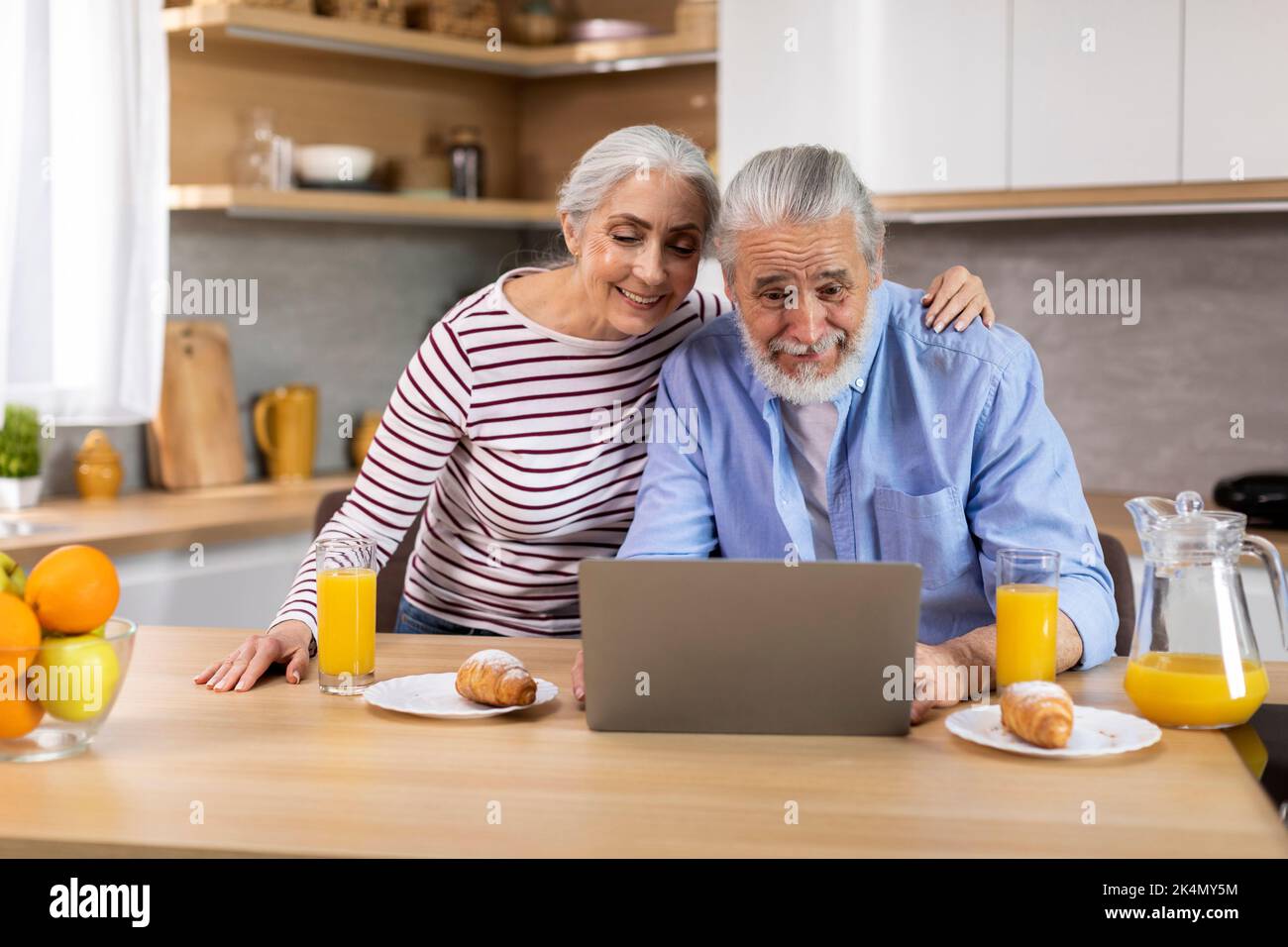 Smiling Elderly Spouses Relaxing With Laptop While Having Breakfast In Kitchen Stock Photo