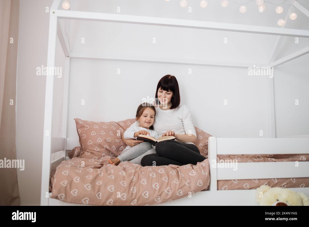 Portrait of cheerful caucasian preschooler girl and her young lady mother reading funny interesting children book together while sitting on bed in bedroom interior. Stock Photo