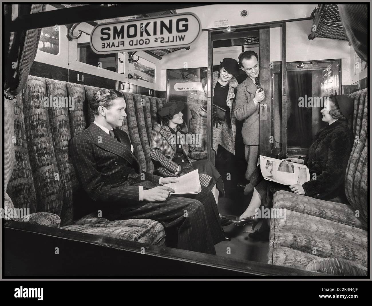 UK British Rail Vintage First Class Rail Compartment with 'SMOKING' sign sponsored by Wills Gold Flake and happy well dressed passengers in a comfortable railway train carriage sponsored smoking compartment,  Archive Retro Britain UK 1936. Stock Photo