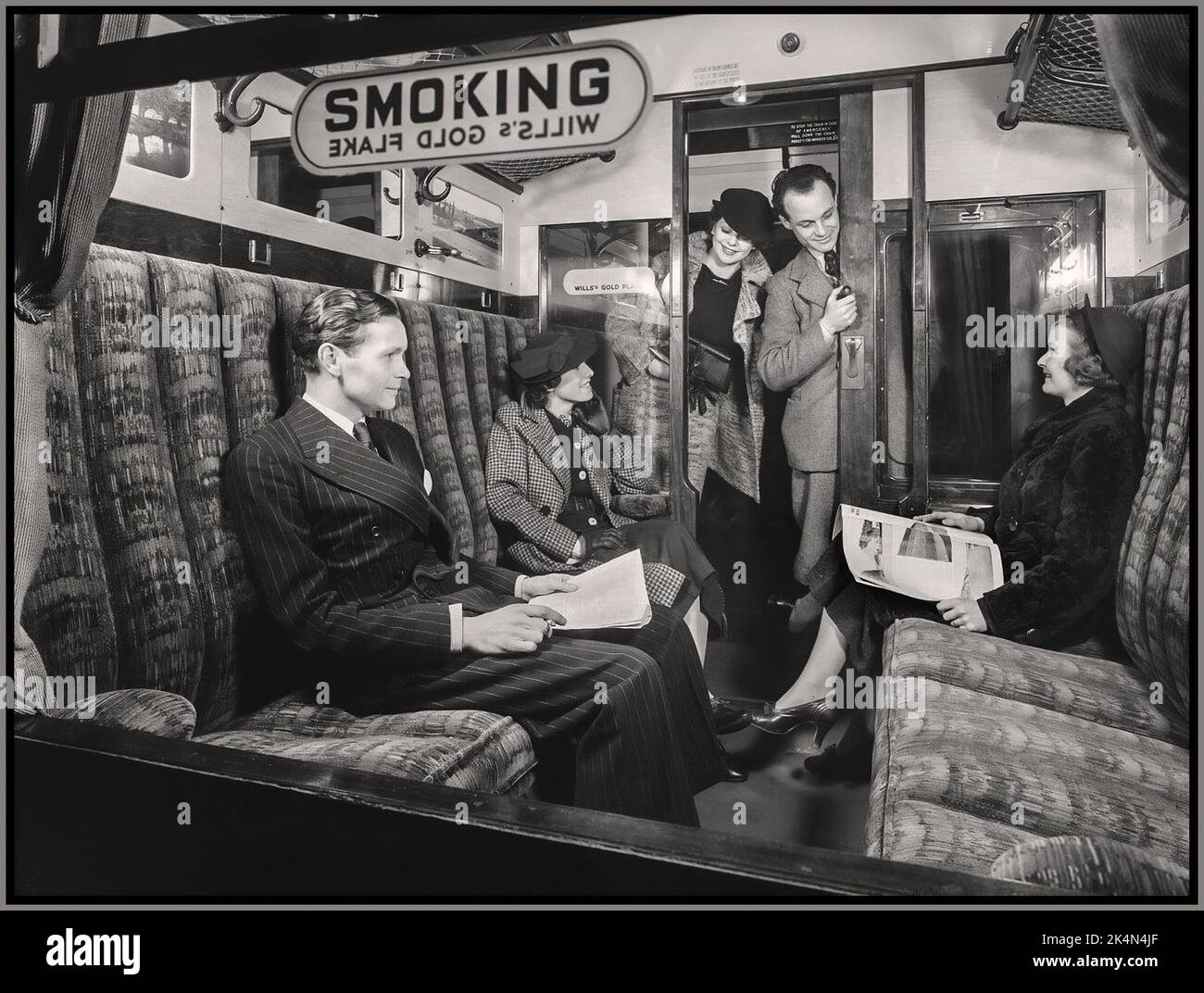 UK British Rail Vintage First Class Rail Compartment with 'SMOKING' sign sponsored by Wills Gold Flake and happy well dressed passengers in a comfortable railway train carriage tobacco sponsored smoking compartment,  Archive Retro Britain UK 1936. Stock Photo