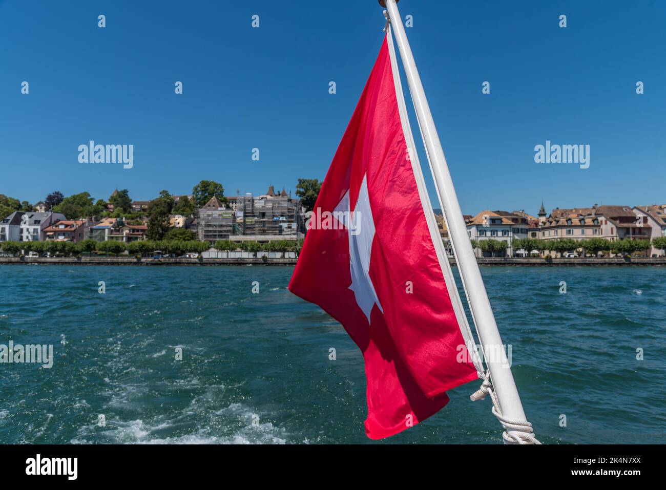Swiss flag fluttering in the wind from the stern of a ship with the wake on Lake Geneva. Lausanne at the background  Stock Photo