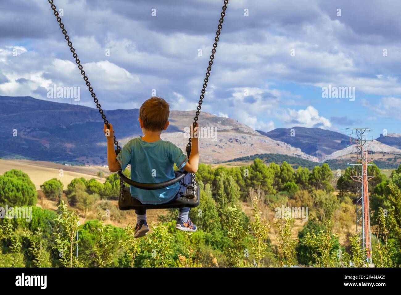 child with unrecognizable back swinging with a cloudy landscape in the background Stock Photo