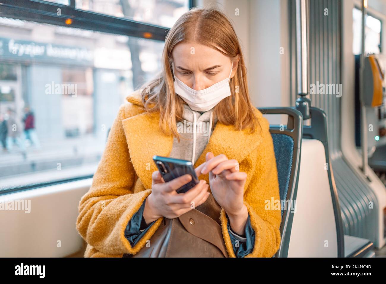 Young woman in medical mask sitting in public bus and reading news in smartphone online during pandemic Covid-19. New normal concept. Stock Photo