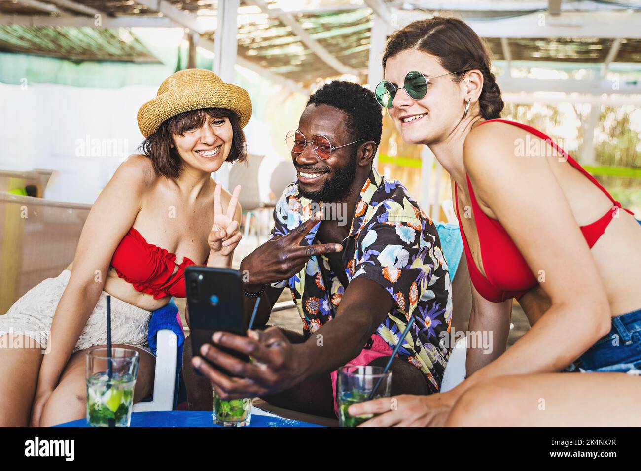 One African guy and two caucasian women having fun enjoying taking selfies at the kiosk bar - people drinking mojito cocktails and shooting self portr Stock Photo