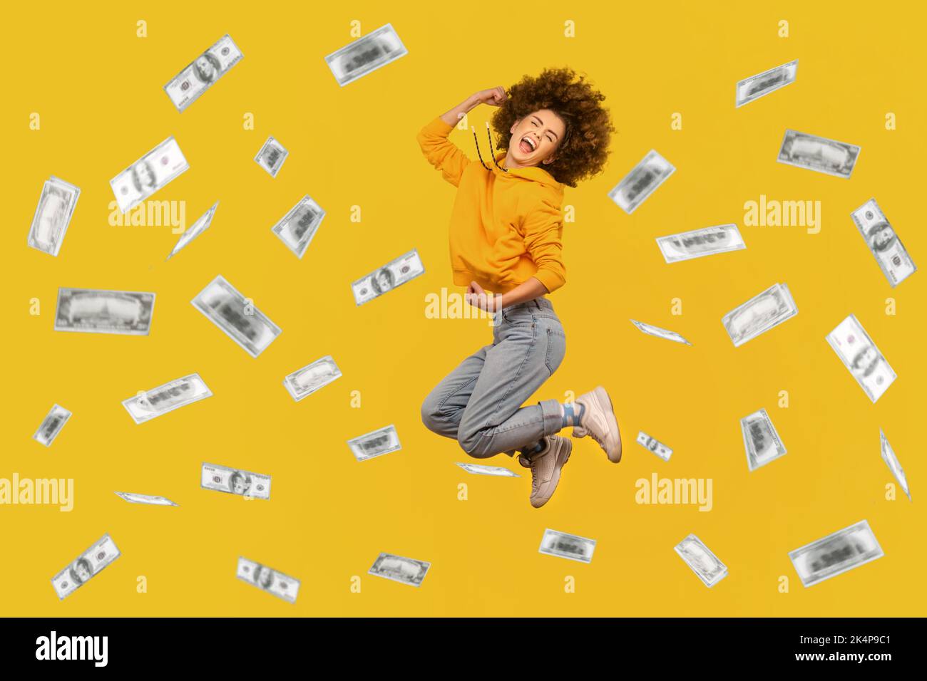 Portrait of successful rich woman with Afro hairstyle jumping in money rain, celebrating her victory, clenched fists and screaming happily. Indoor studio shot isolated on yellow background. Stock Photo