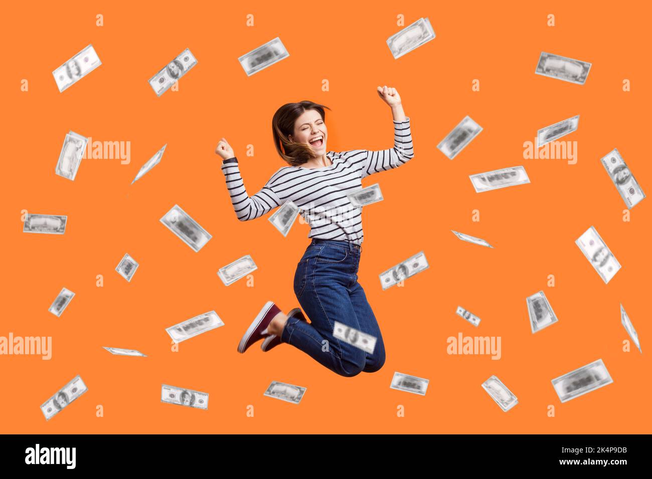 Portrait of satisfied excited woman jumping with clenched fist, celebrating winning lottery, flying in money rain, dollars falling. Indoor studio shot isolated on orange background. Stock Photo