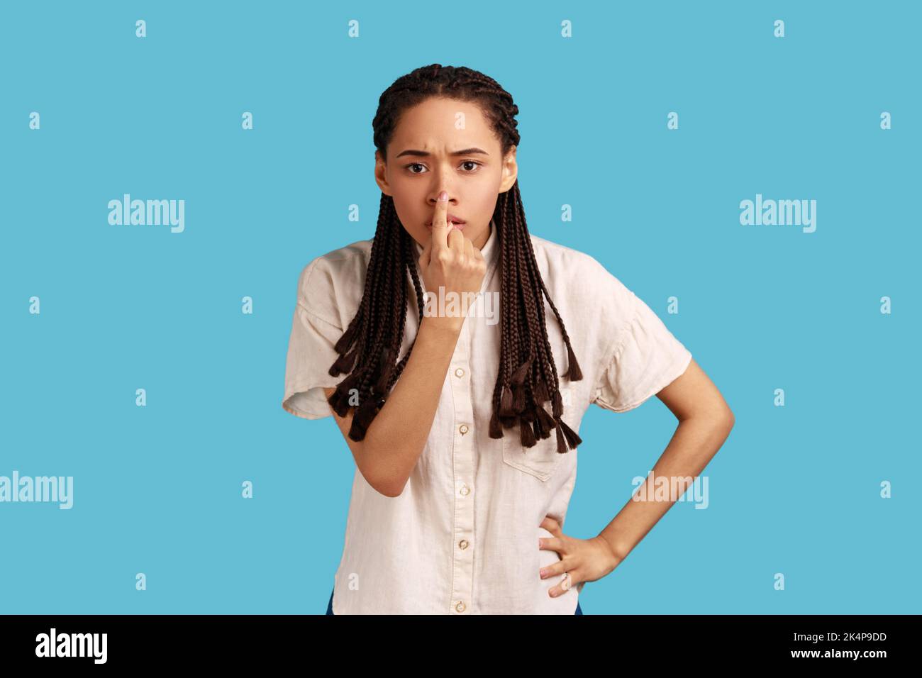 You are liar. Portrait of serious woman with black dreadlocks standing with finger on her nose and showing lie gesture, wearing white shirt. Indoor studio shot isolated on blue background. Stock Photo