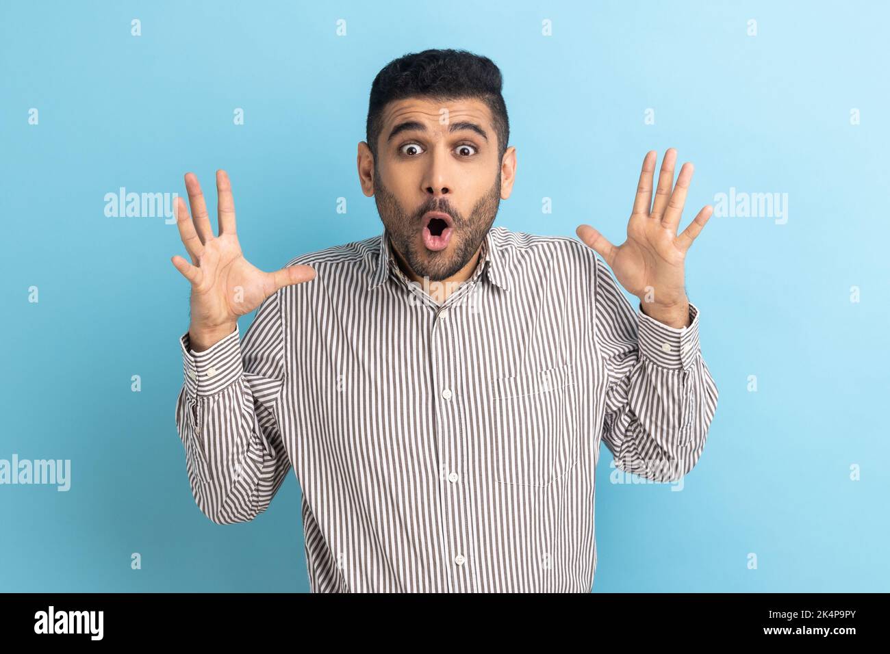 Portrait of surprised astonished bearded businessman standing with open mouth and raised arms, being impressed of shocking news, wearing striped shirt. Indoor studio shot isolated on blue background. Stock Photo