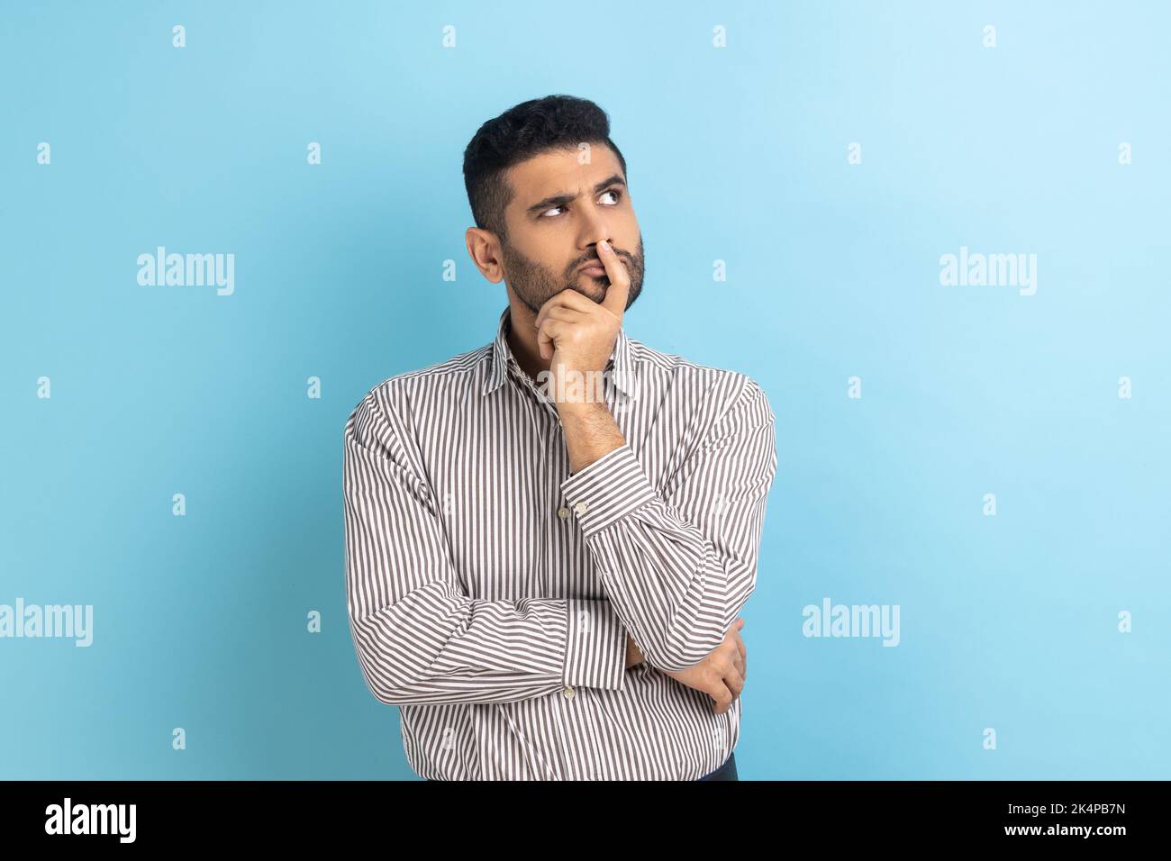 Portrait of thoughtful handsome bearded businessman holding his chin and pondering idea, confused not sure about solution, wearing striped shirt. Indoor studio shot isolated on blue background. Stock Photo