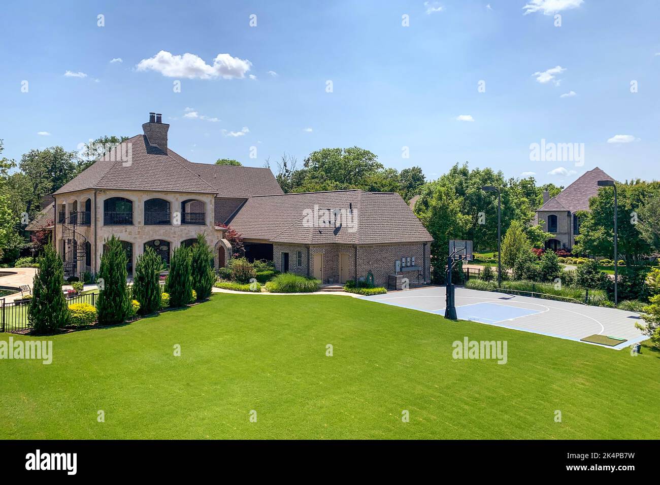 Outdoor shot of luxury house in suburban neighborhood with trees, bushes, lawn and basketball court on the backyard, blue sky background. Real estate concept. Stock Photo