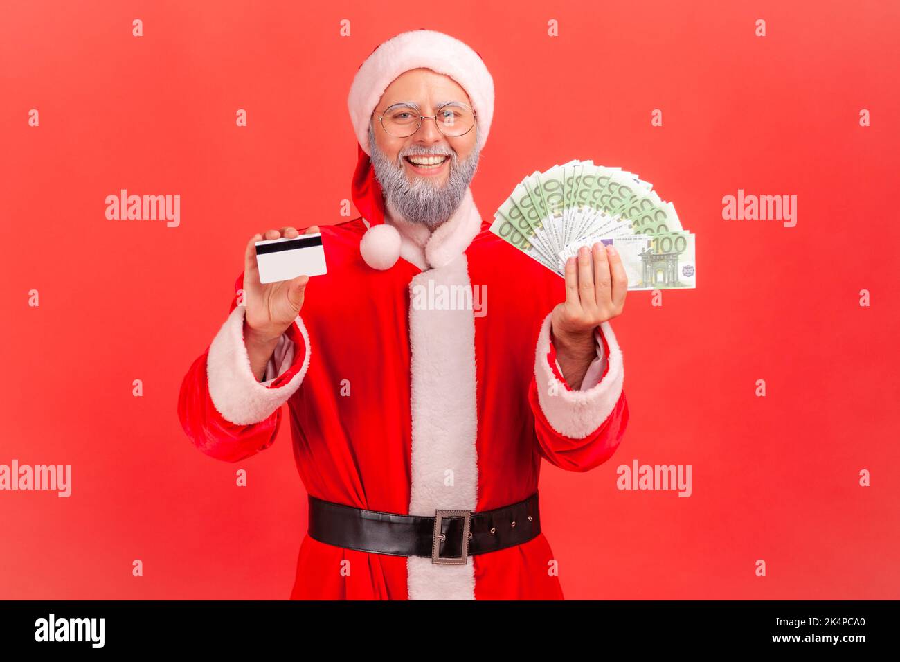 Happy smiling satisfied elderly man with gray beard wearing santa claus costume holding showing fan of euro banknotes and credit card in hands. Indoor studio shot isolated on red background. Stock Photo