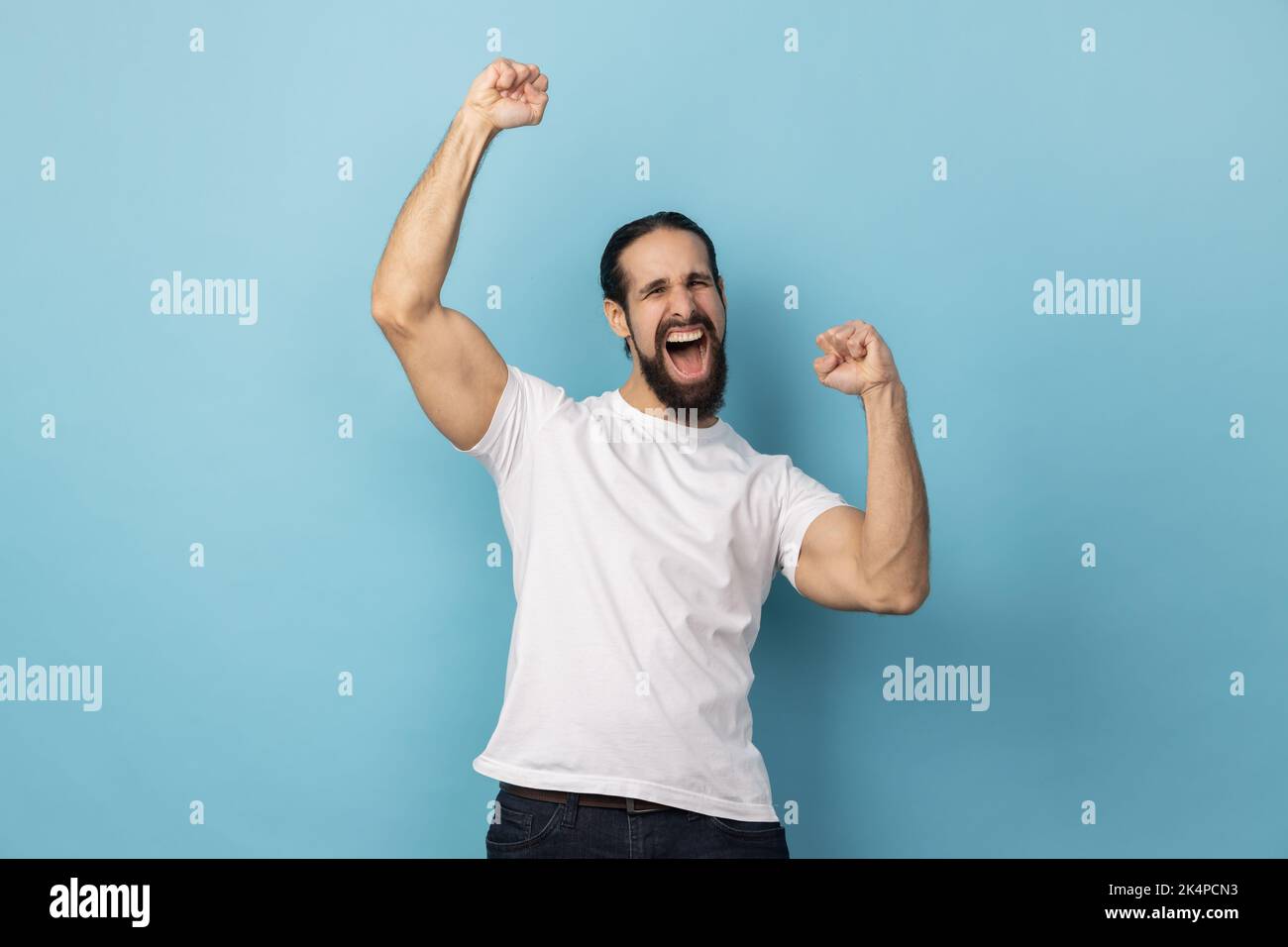 Portrait of excited man with beard wearing white T-shirt expressing winning gesture with raised fists and screaming, celebrating victory. Indoor studio shot isolated on blue background. Stock Photo