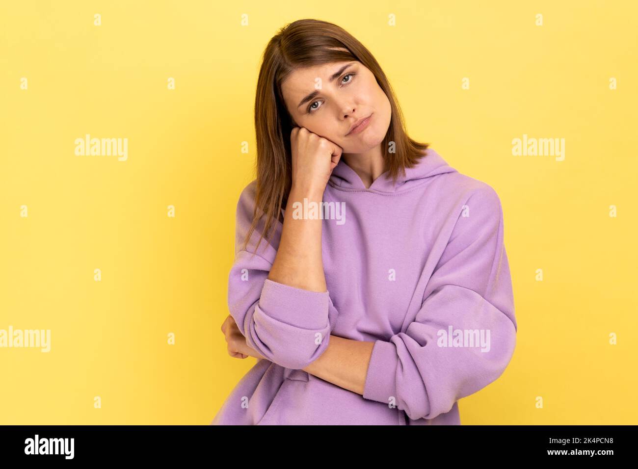 Portrait of bored exhausted woman leaning on hand and looking at camera with disinterest, tired of boring sad story, wearing purple hoodie. Indoor studio shot isolated on yellow background. Stock Photo
