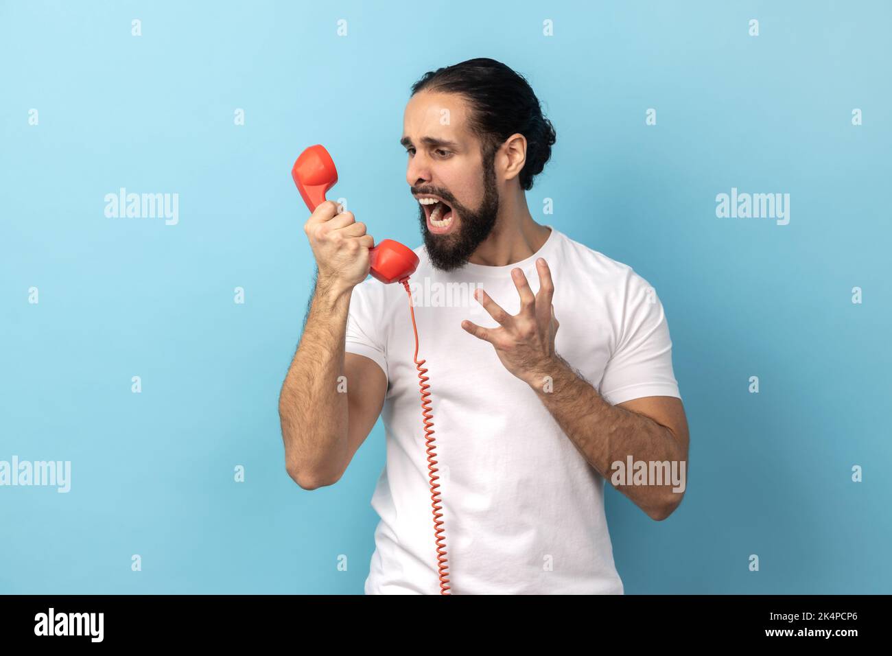 Portrait of angry man with beard wearing white T-shirt screaming and yelling talking retro landline phone, complaining on connection quality. Indoor studio shot isolated on blue background. Stock Photo