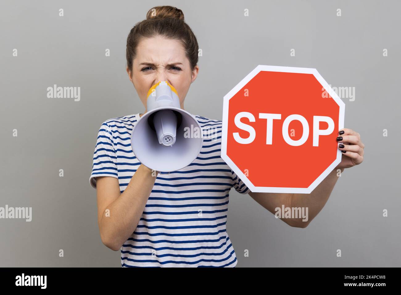 Portrait of excited woman wearing striped T-shirt looking at camera, holding red stop sign and screaming in megaphone in her hands. Indoor studio shot isolated on gray background. Stock Photo