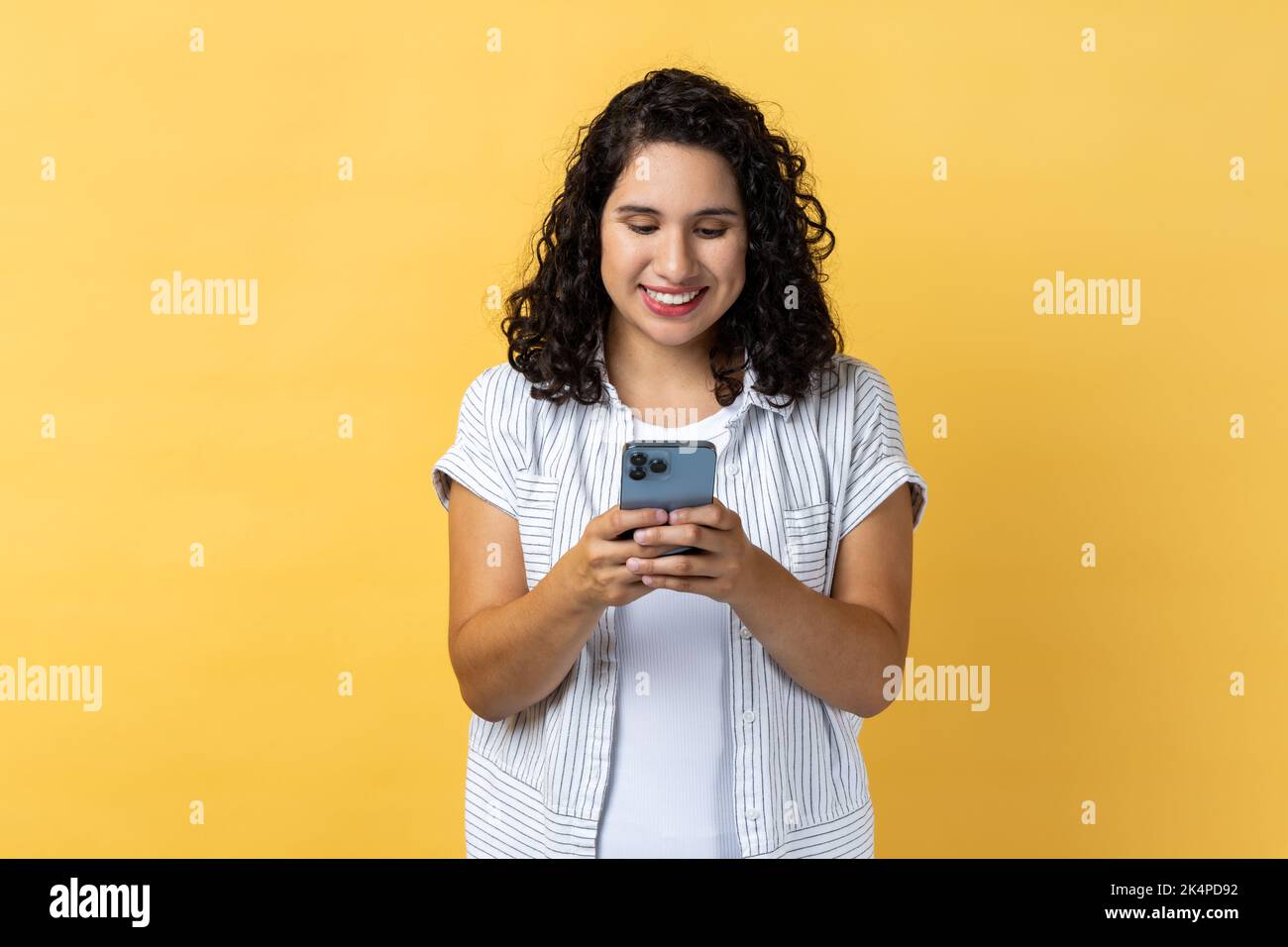 Portrait of smiling satisfied young adult woman with dark wavy hair using mobile phone with happy expression, checking social networks. Indoor studio shot isolated on yellow background. Stock Photo