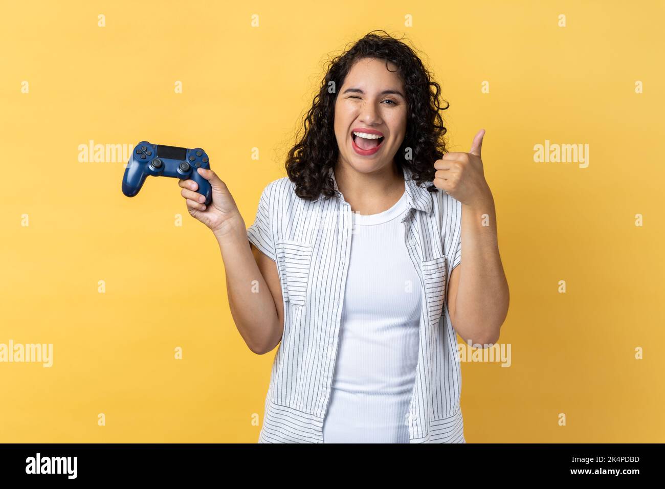 Portrait of joyful beautiful woman with dark wavy hair holding joystick in hands and showing thumb up, likes playing games on joypad. Indoor studio shot isolated on yellow background. Stock Photo