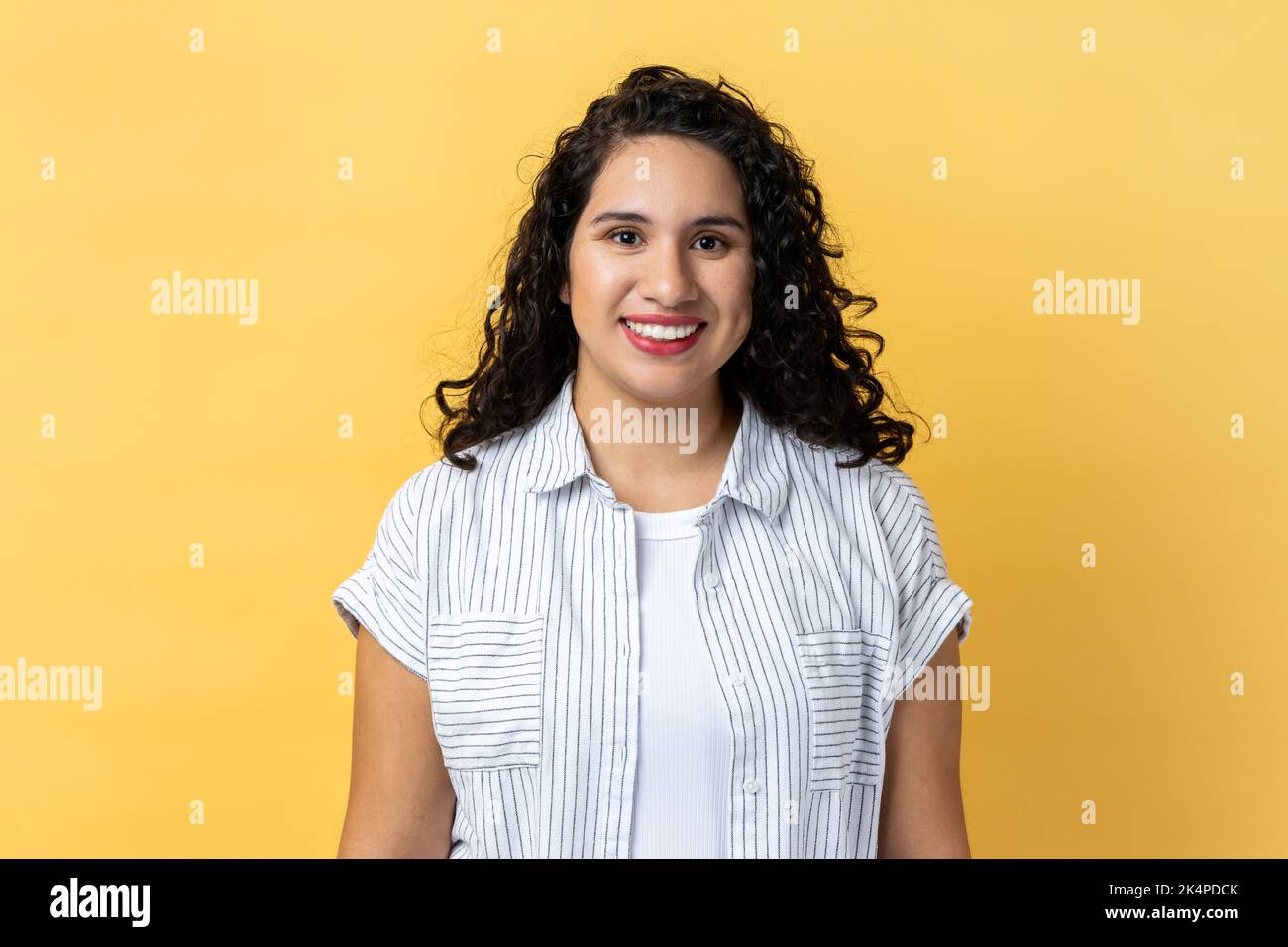 Portrait of cheerful positive woman with dark wavy hair looking at camera with toothy smile, expressing happiness, being in good mood. Indoor studio shot isolated on yellow background. Stock Photo