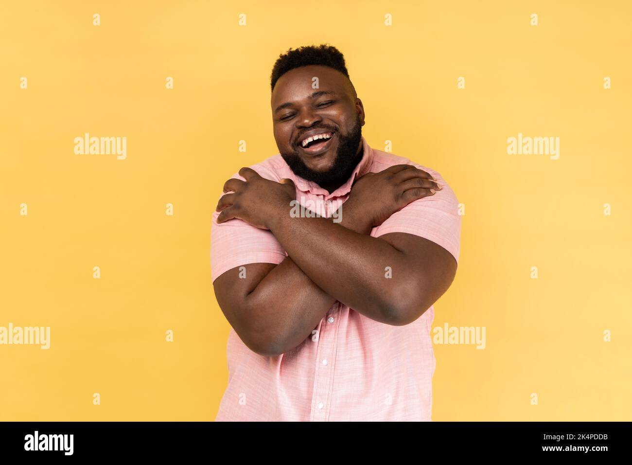 Portrait of selfish narcissistic man wearing pink shirt embracing himself and smiling with expression of great ego, pleasure and self-esteem. Indoor studio shot isolated on yellow background. Stock Photo