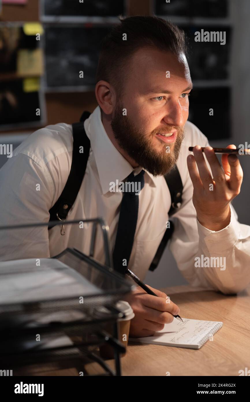 Portrait of an investigator working in the office at desk, detective dictating to voice assistant on a mobile phone, searching for information by Stock Photo