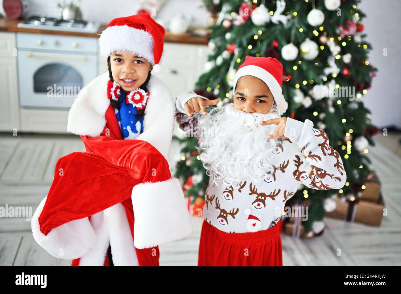 African American children - brother and sister in a large size santa costume with a false beard play and grimace against the backdrop of the Christmas Stock Photo