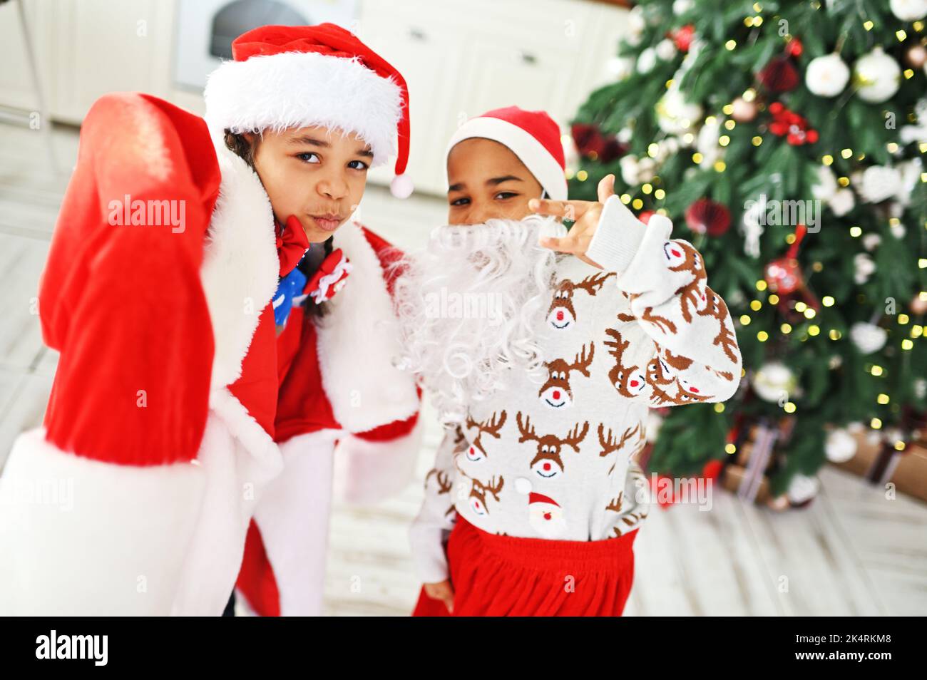 children - brother and sister in a large size santa costume with a false beard play and grimace against the backdrop of the Christmas tree. Stock Photo