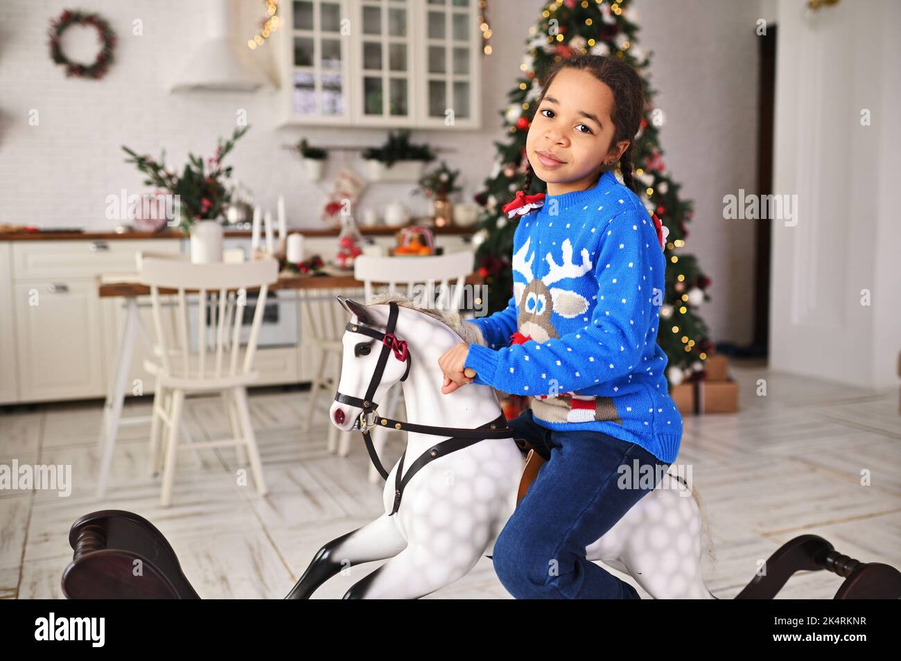 little African-American girl in a blue Christmas sweater smiles while sitting on a toy rocking horse against the background of a Christmas tree and a Stock Photo