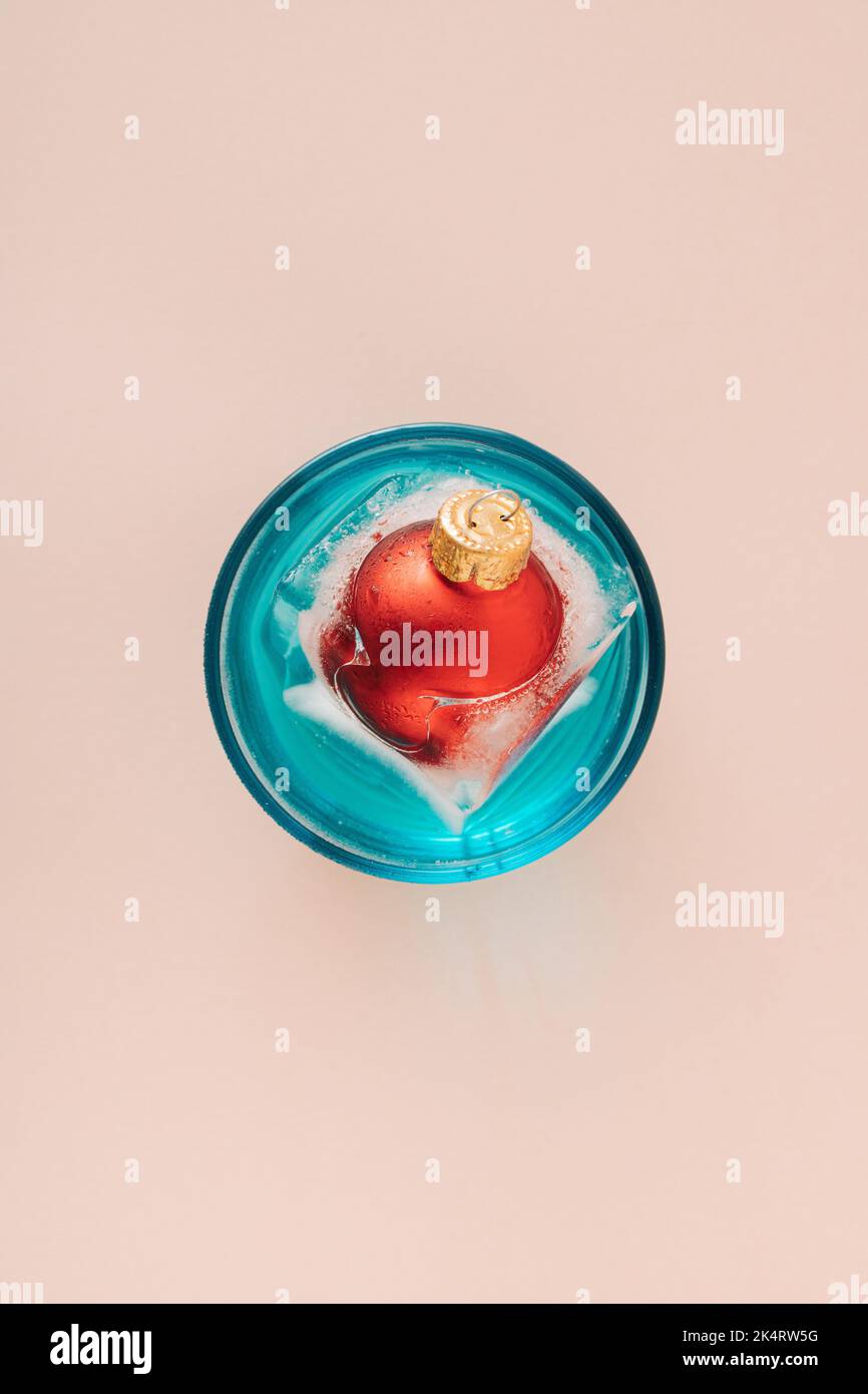 Red Christmas bauble in melting ice cube floating in blue drinking glass. New year greetings. Minimal concept. Stock Photo