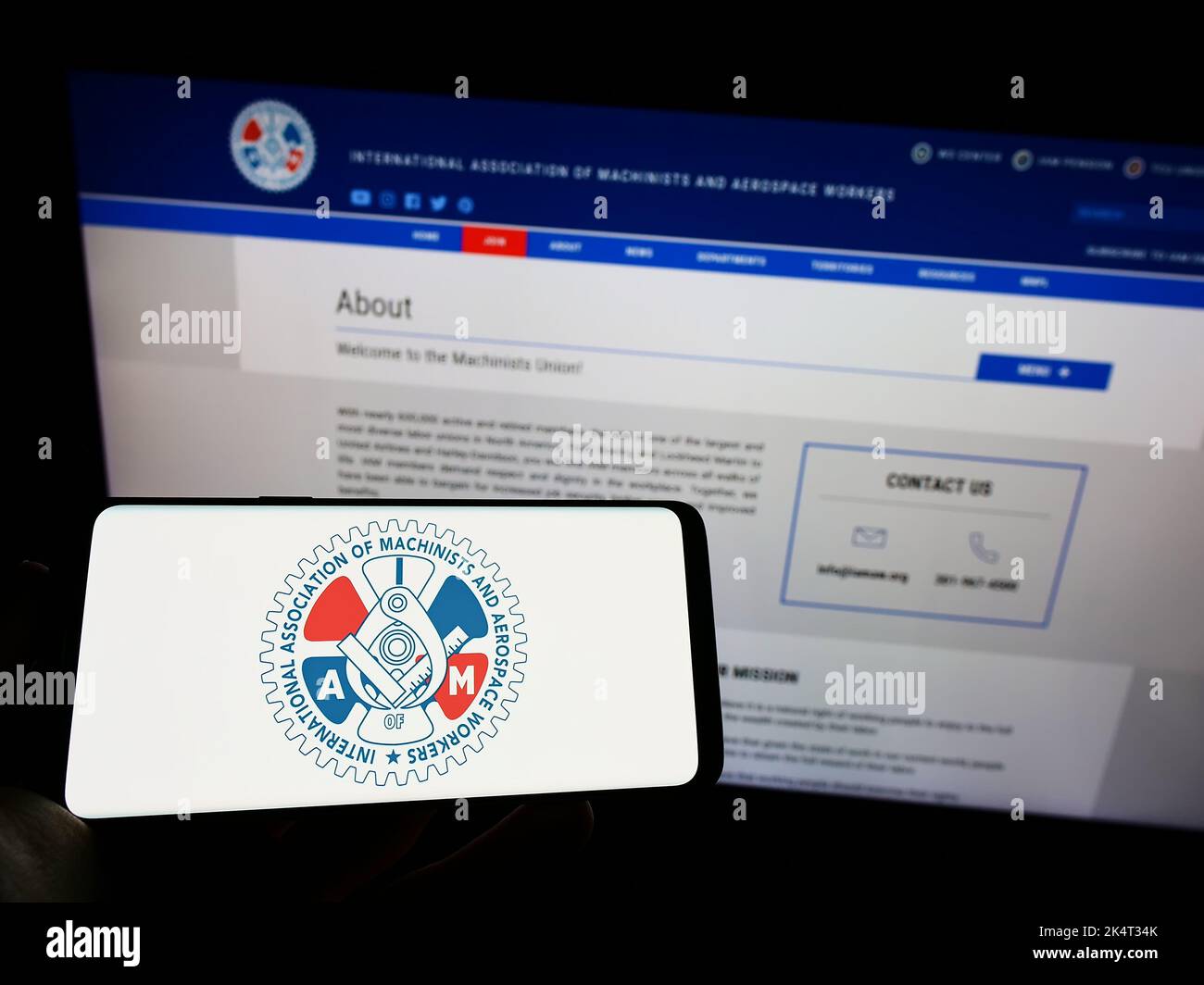 Person holding mobile phone with logo of American-Canadian trade union IAM on screen in front of web page. Focus on phone display. Stock Photo