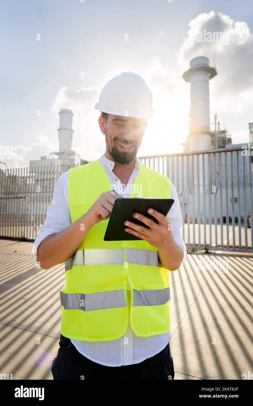 Vertical photo of smiley caucasian engineer using digital tablet to do inspections at energy plant. Stock Photo