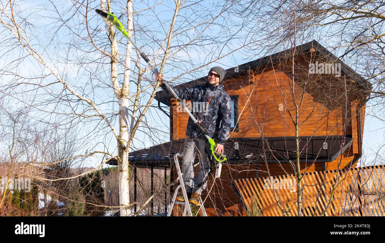 A gardener prunes a tree with an electric brush cutter. A man is standing on a stepladder and using an electric chain garden pruner with a telescopic Stock Photo