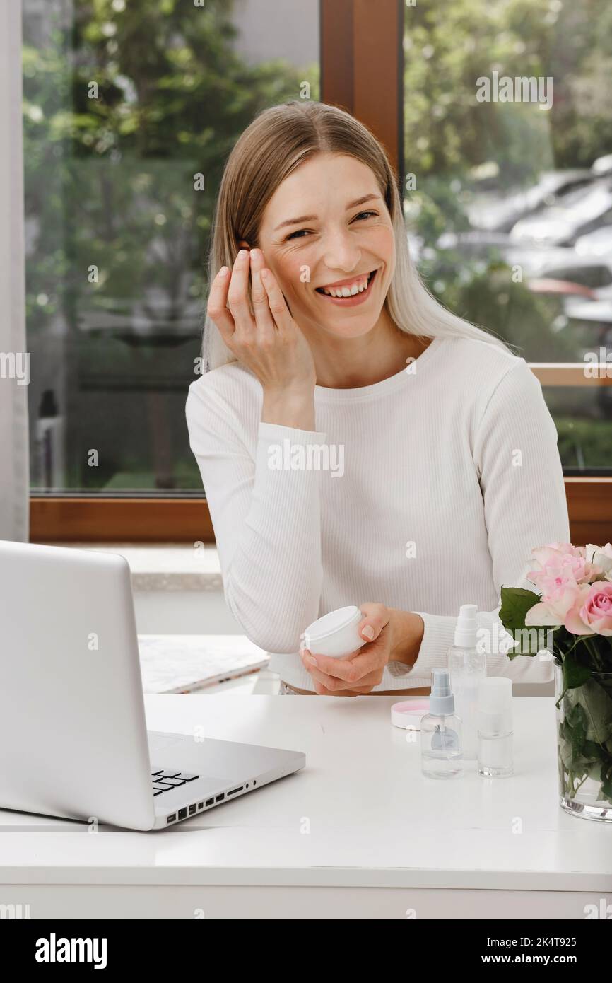 Applying Cream on beauty blogger face near laptop. Woman making online video content, live broadcast review on cosmetic products. Cosmetology course Stock Photo