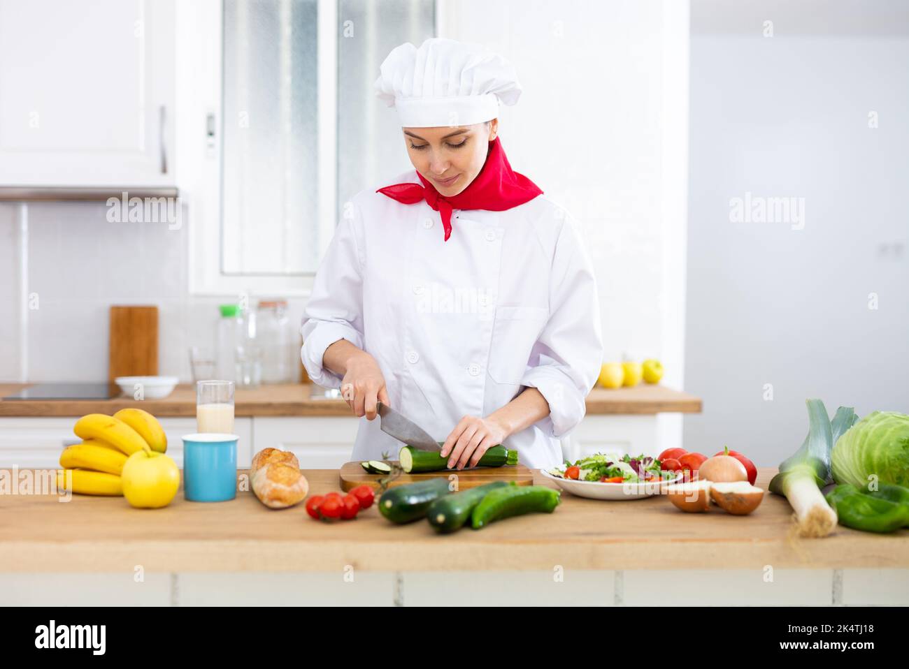 Smiling female chef in white uniform preparing vegetable salad in private kitchen Stock Photo