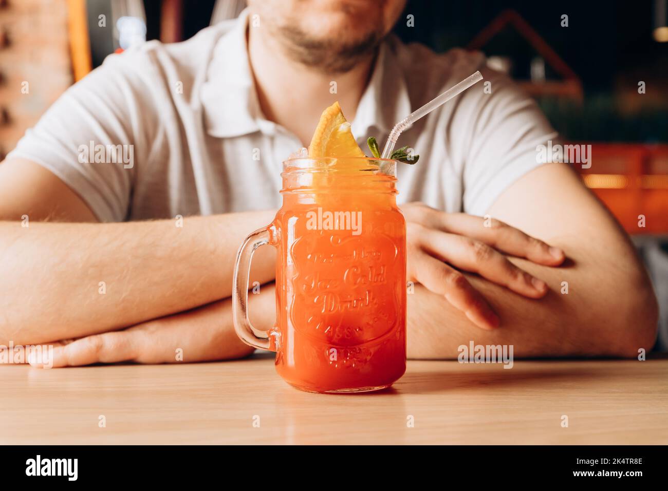 Young caucasian man and Red Strawberry lemonade with ice,  cocktail tube and orange slice in glass with handle on wooden table. Male hands and cocktai Stock Photo