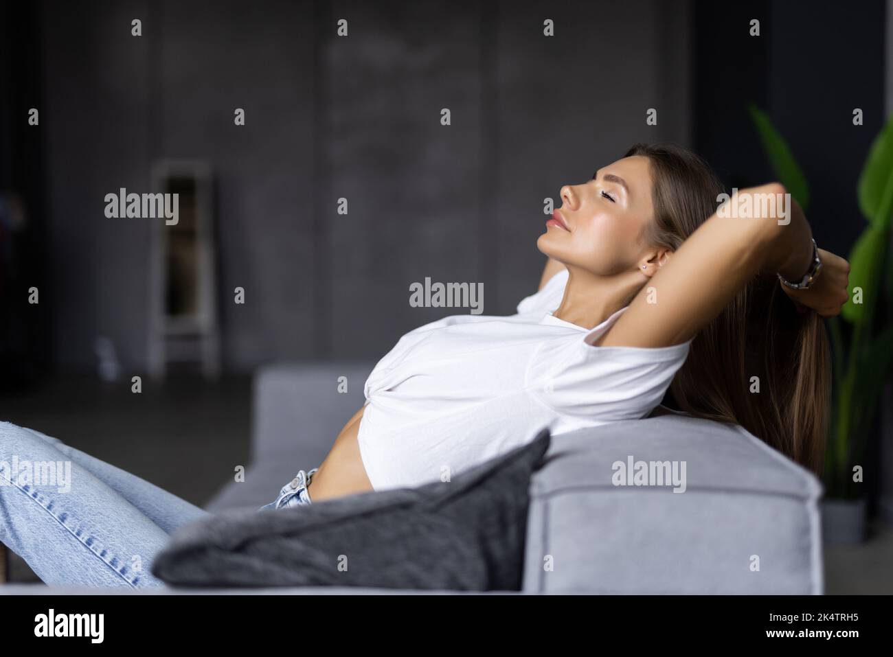 Calm serene young woman relaxing on comfortable couch at home with closed eyes and hands behind head, satisfied girl stretching on sofa, daydreaming a Stock Photo