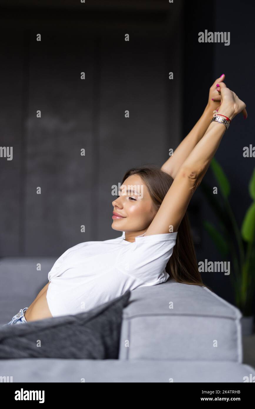 Calm serene young woman relaxing on comfortable couch at home with closed eyes and hands behind head, satisfied girl stretching on sofa, daydreaming a Stock Photo