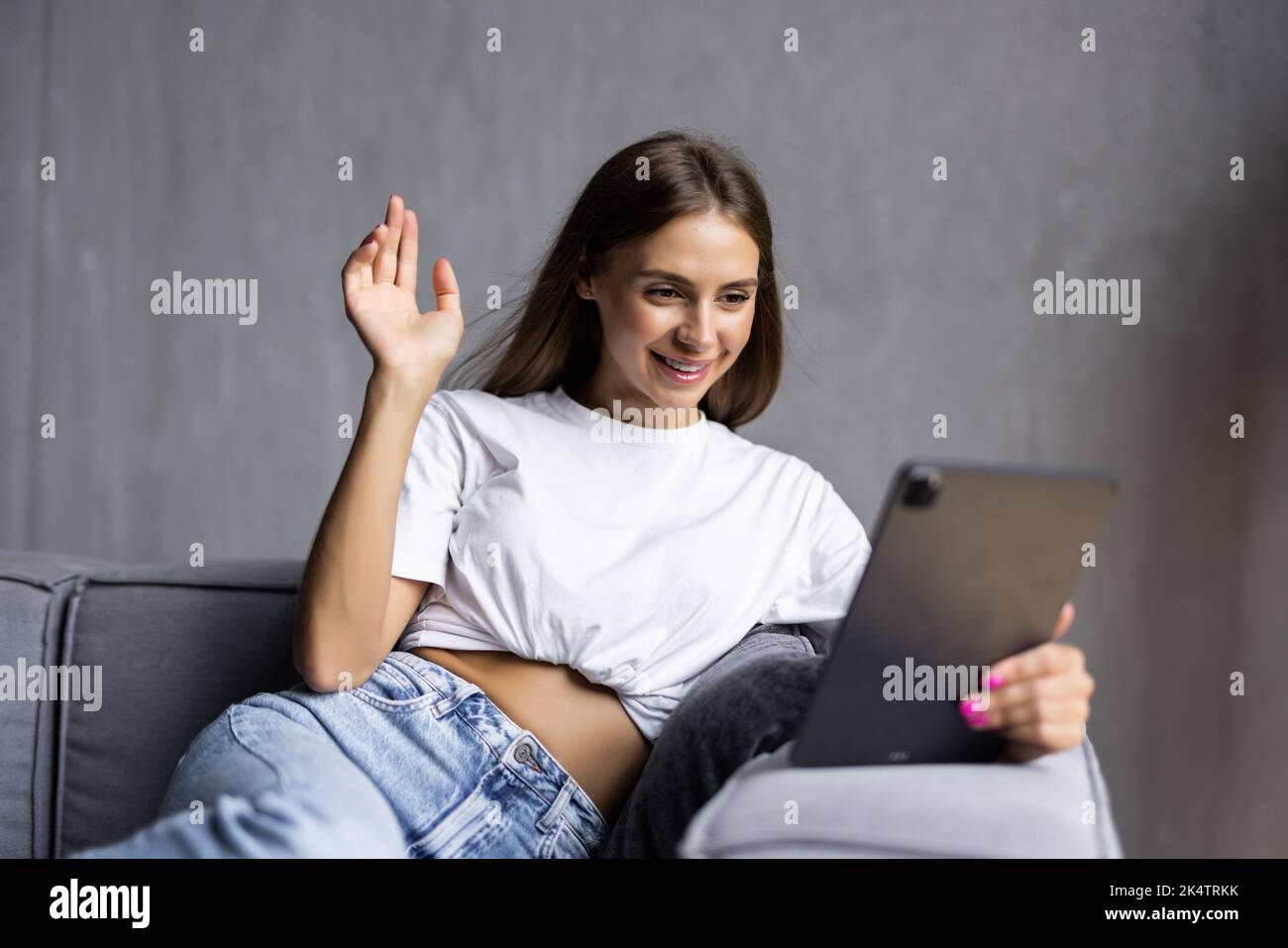 Gorgeous smiling caucasian woman sitting on sofa and using tablet for video call and waving. Living room interior. Stock Photo