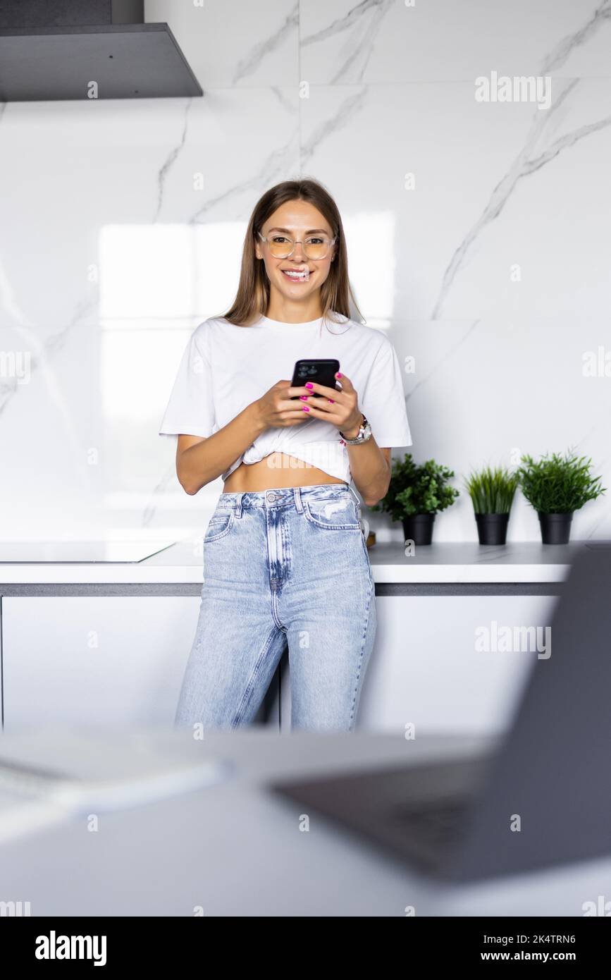 Pretty young woman standing in the kitchen at daytime and using her smartphone Stock Photo