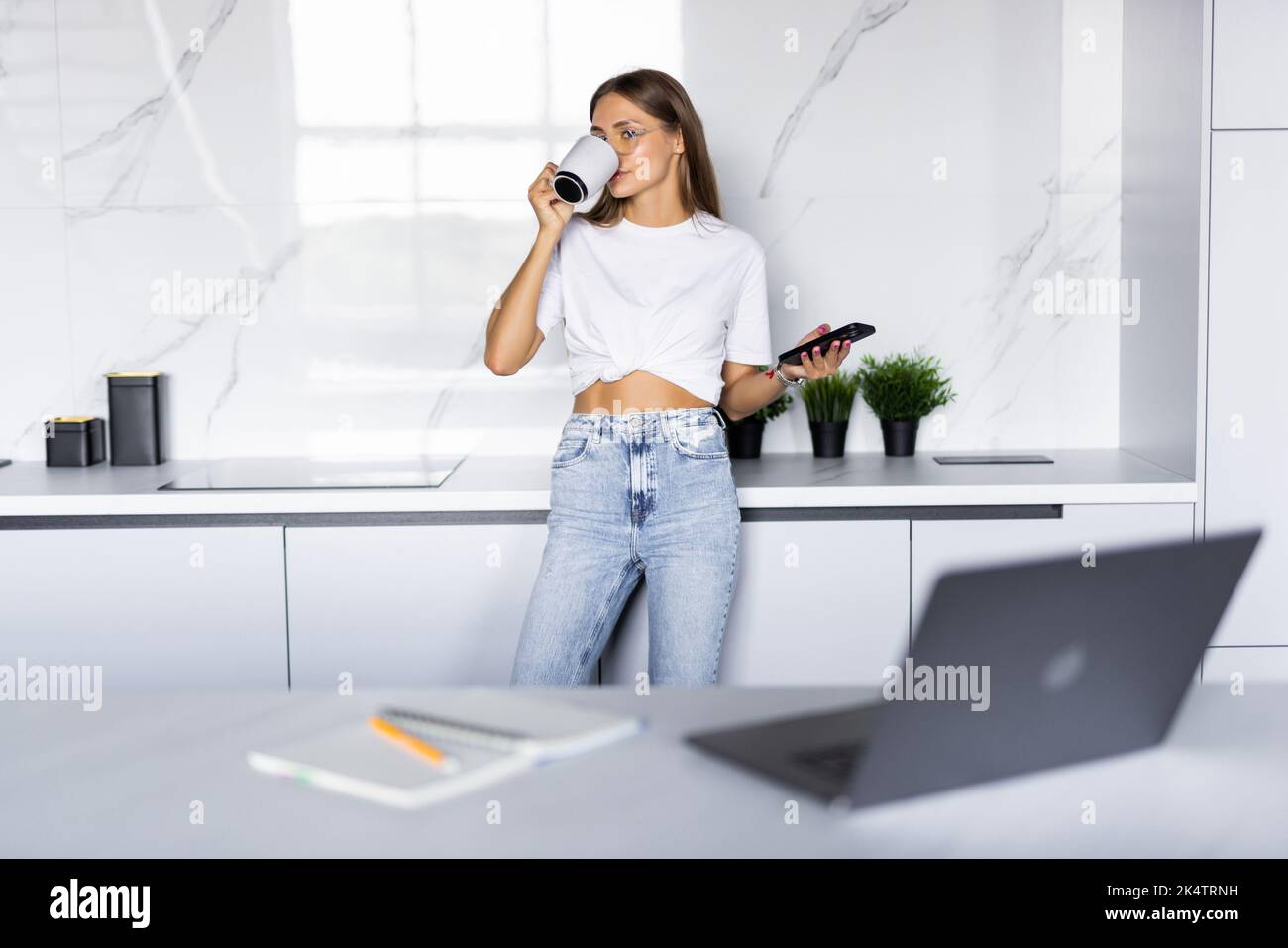 Young woman using smartphone leaning at kitchen table with coffee mug and organizer in a modern home. Smiling woman reading phone message. Brunette ha Stock Photo